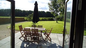 Eine Terrasse im Freien mit Tisch und Stühlen, einem schwarzen Sonnenschirm und Blick auf eine Rasenfläche mit Bäumen.