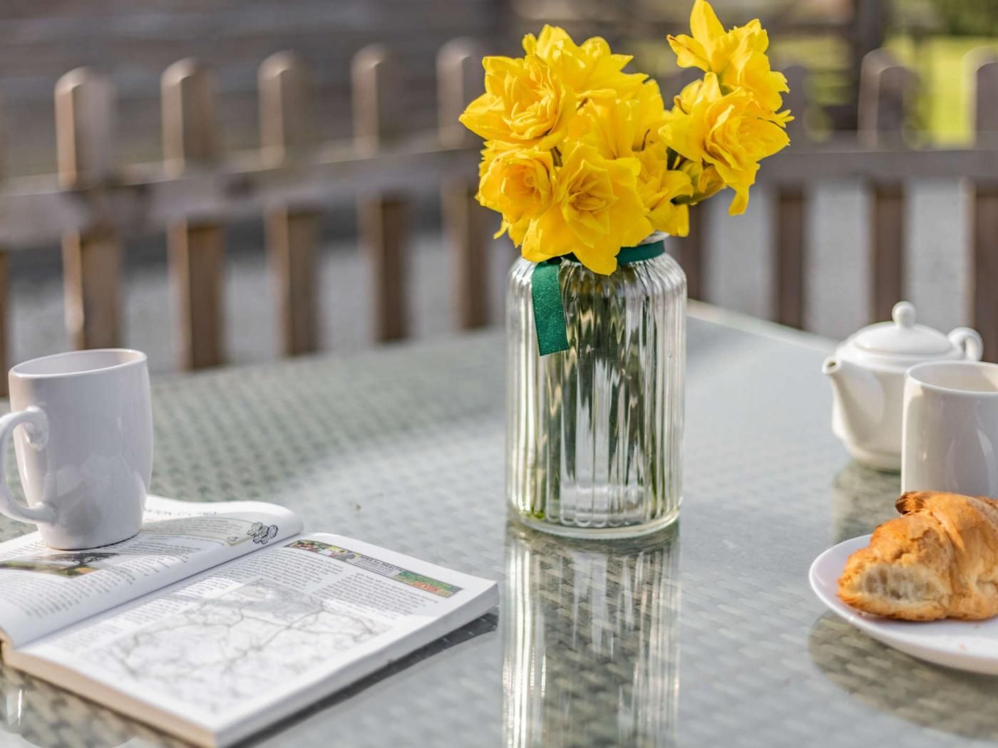A table with a clear glass vase containing yellow flowers, a white teapot, a white cup, a plate with a pastry, and a menu.