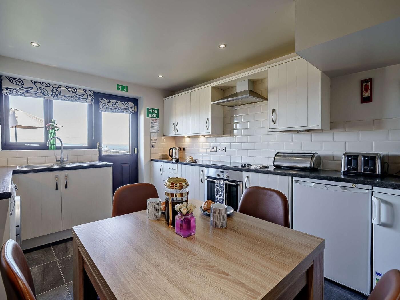 A modern kitchen with white cabinets, a wooden dining table, and a view through the window.