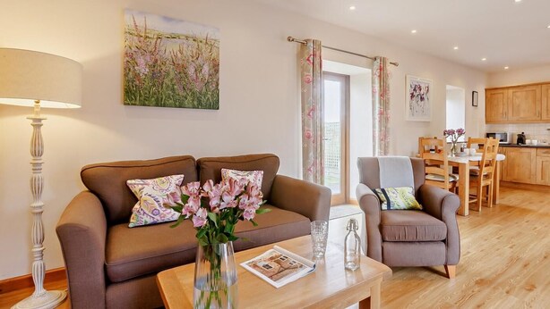 Ein Wohnzimmer mit einer Couch, einem Couchtisch und einem Blumenstrauß. Es gibt einen Essbereich mit Tisch und Stühlen, und im Hintergrund eine Küche.
