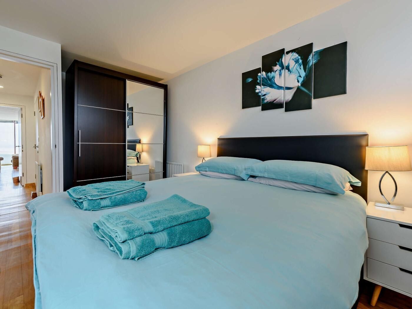 A bedroom with a bed, two bedside tables, a nightstand, a wardrobe, and artwork on the wall.