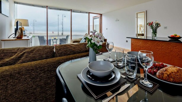 Et moderne spiseområde med havudsigt, en sofa, et bord dækket til to og en køkkenø med en skål med frugt.