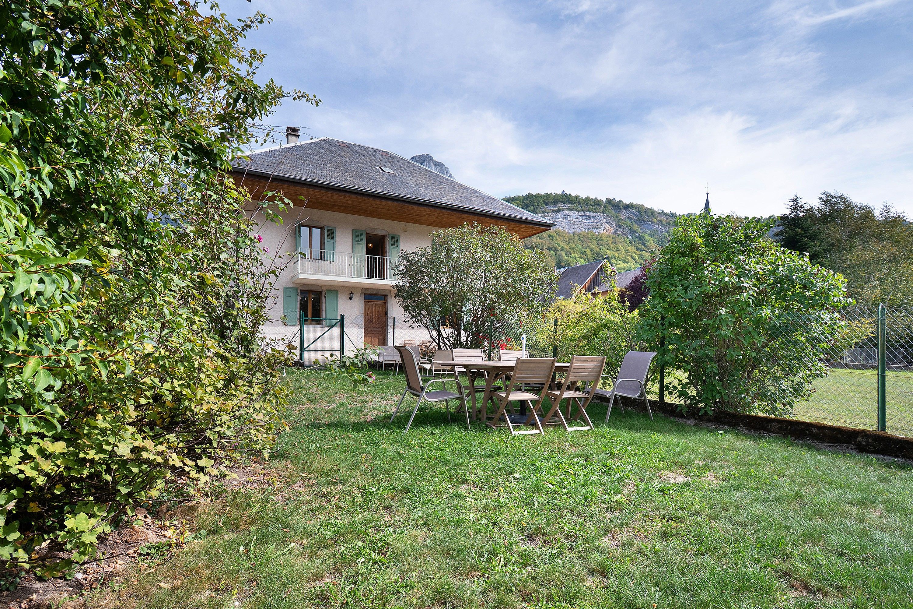 Une maison dotée d’un balcon, entourée de verdure, avec une montagne en toile de fond.