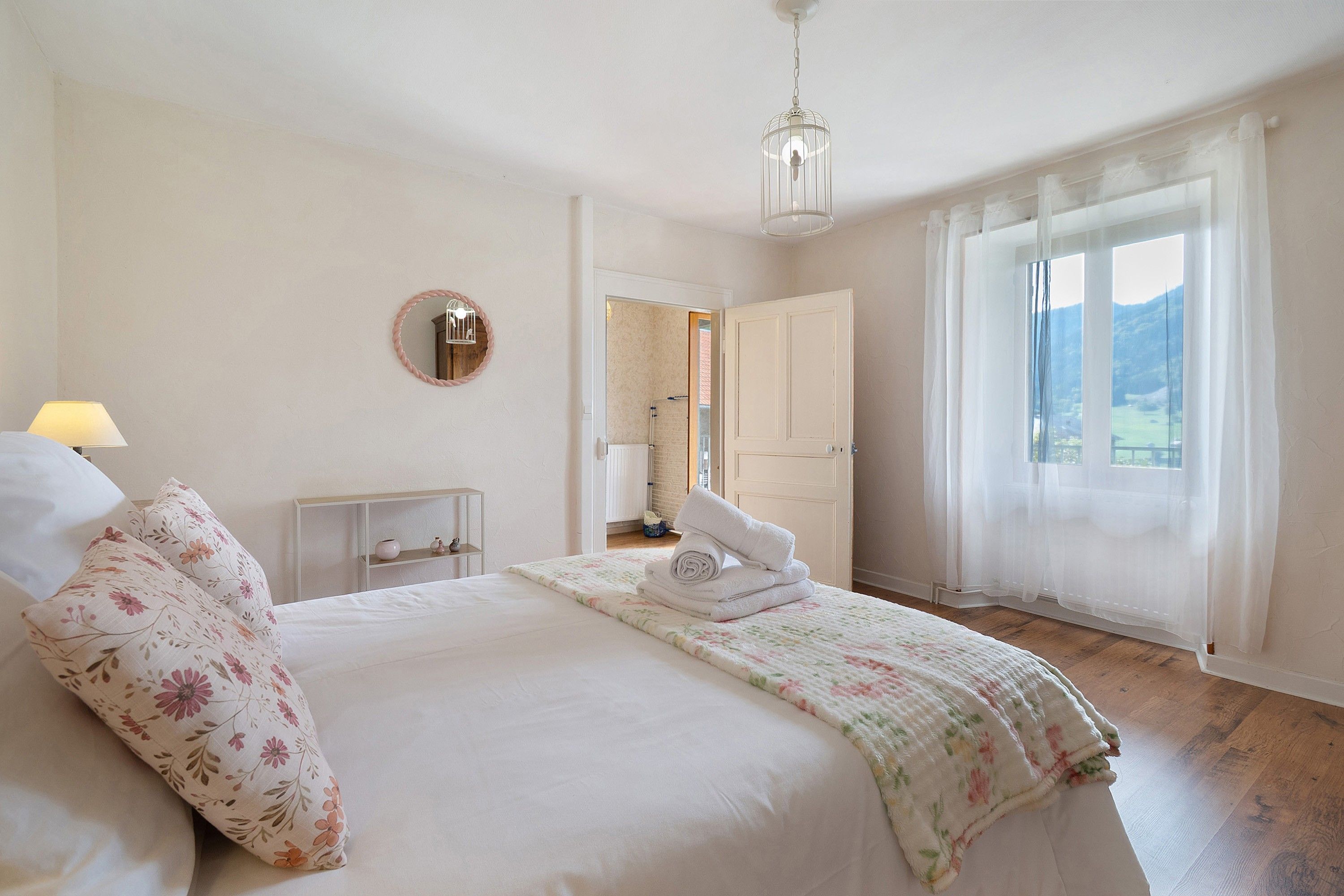 A bedroom with a bed, a bedside table, a lamp, a mirror, and a view of the outdoors through a window.