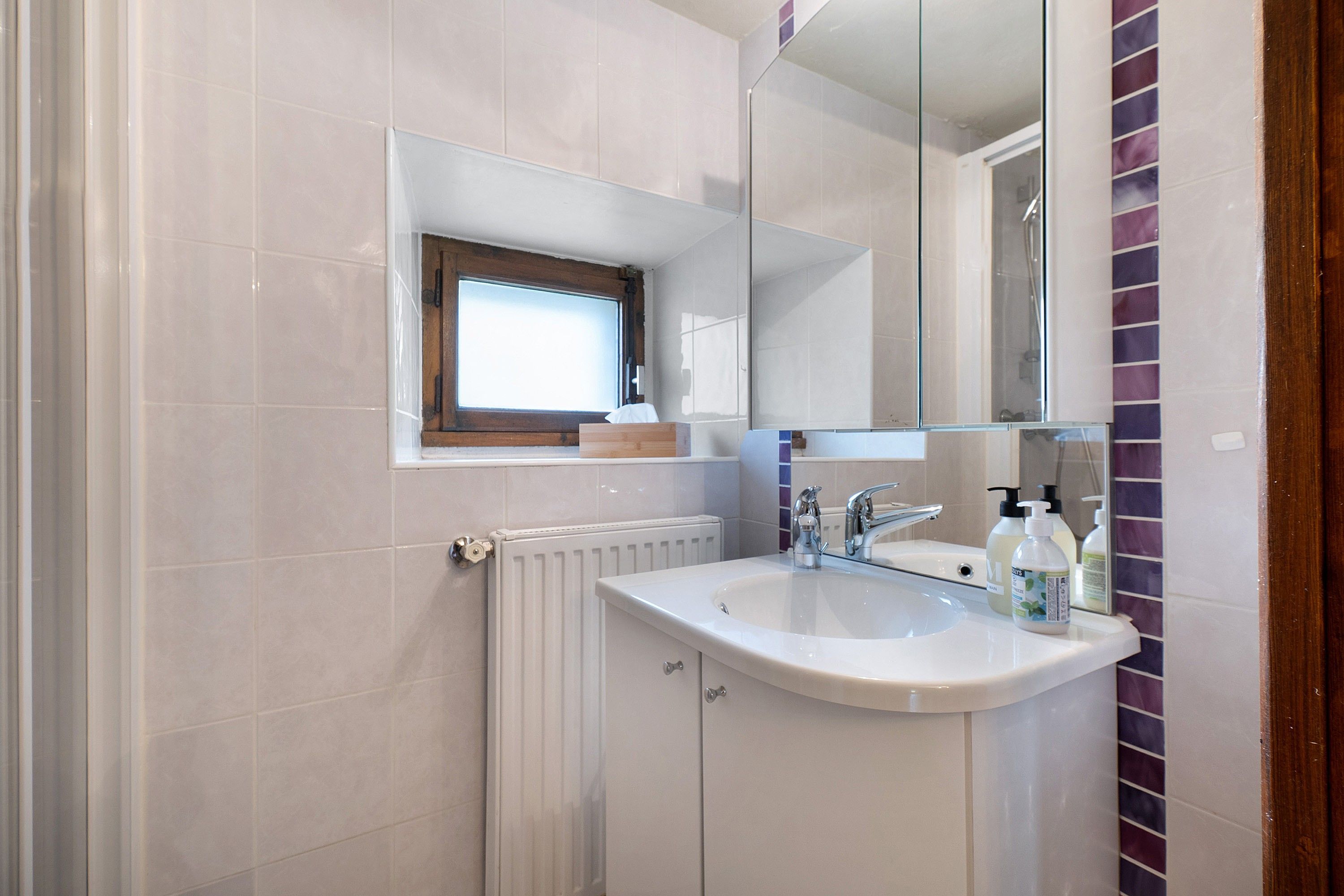 Un bagno con un lavandino bianco, uno specchio e una finestra sopra il lavandino.