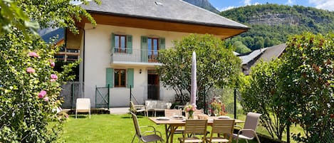 A house with a balcony, a dining area with chairs and a table, and a mountain in the background.