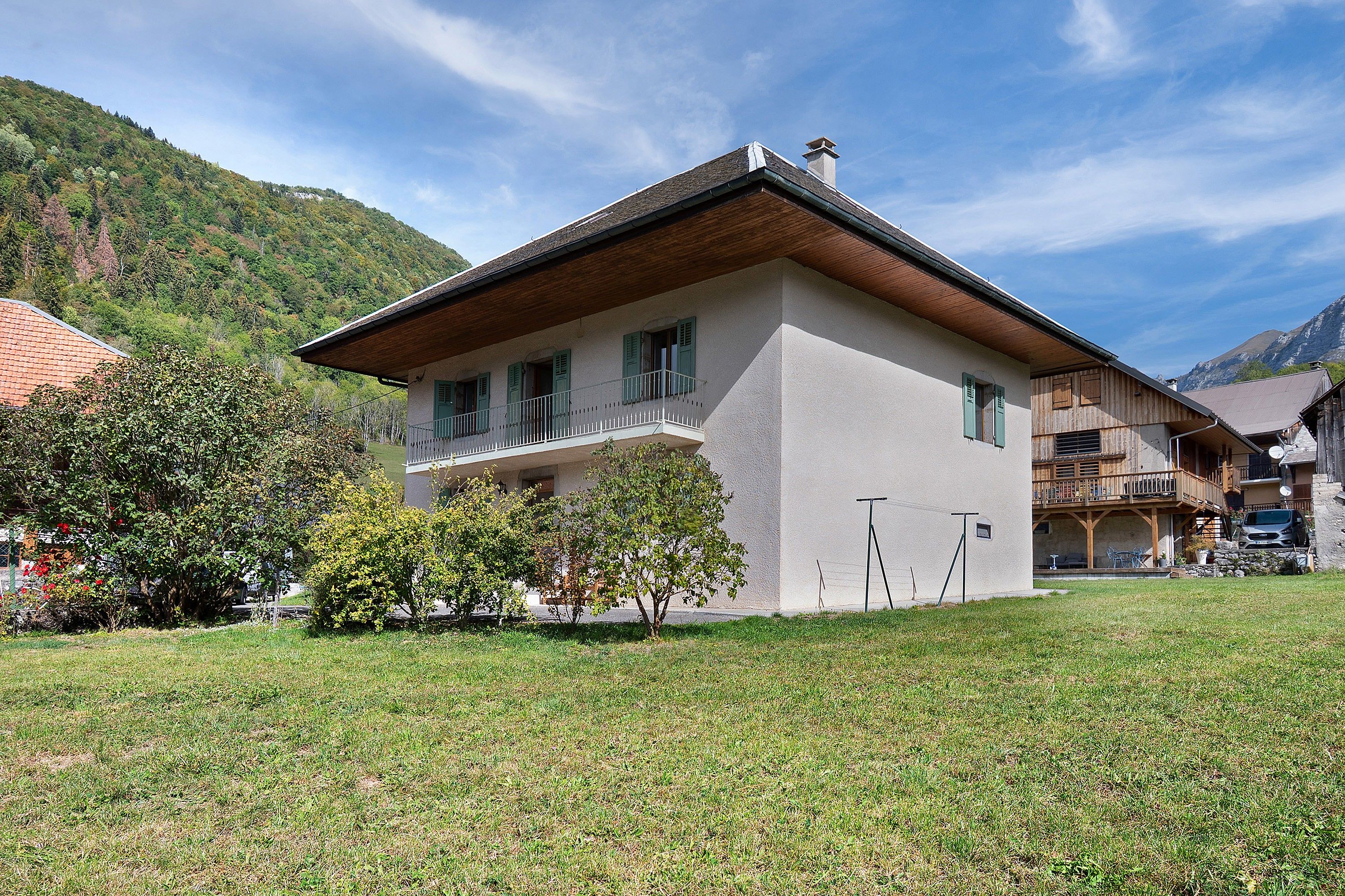 Eine zweigeschossige Chalet mit Balkon, umgeben von einer Rasenfläche und Bäumen, vor einer Bergkulisse gelegen.