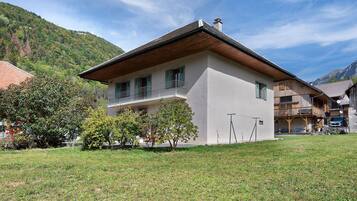 Eine zweigeschossige Chalet mit Balkon, umgeben von einer Rasenfläche und Bäumen, vor einer Bergkulisse gelegen.