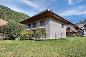 Un chalet à deux étages doté d’un balcon, entouré d’une pelouse verdoyante et d’arbres, avec un paysage montagneux en toile de fond.