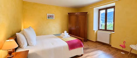 A bedroom with a bed, wooden wardrobe, bedside table, lamp, and a window with a view.
