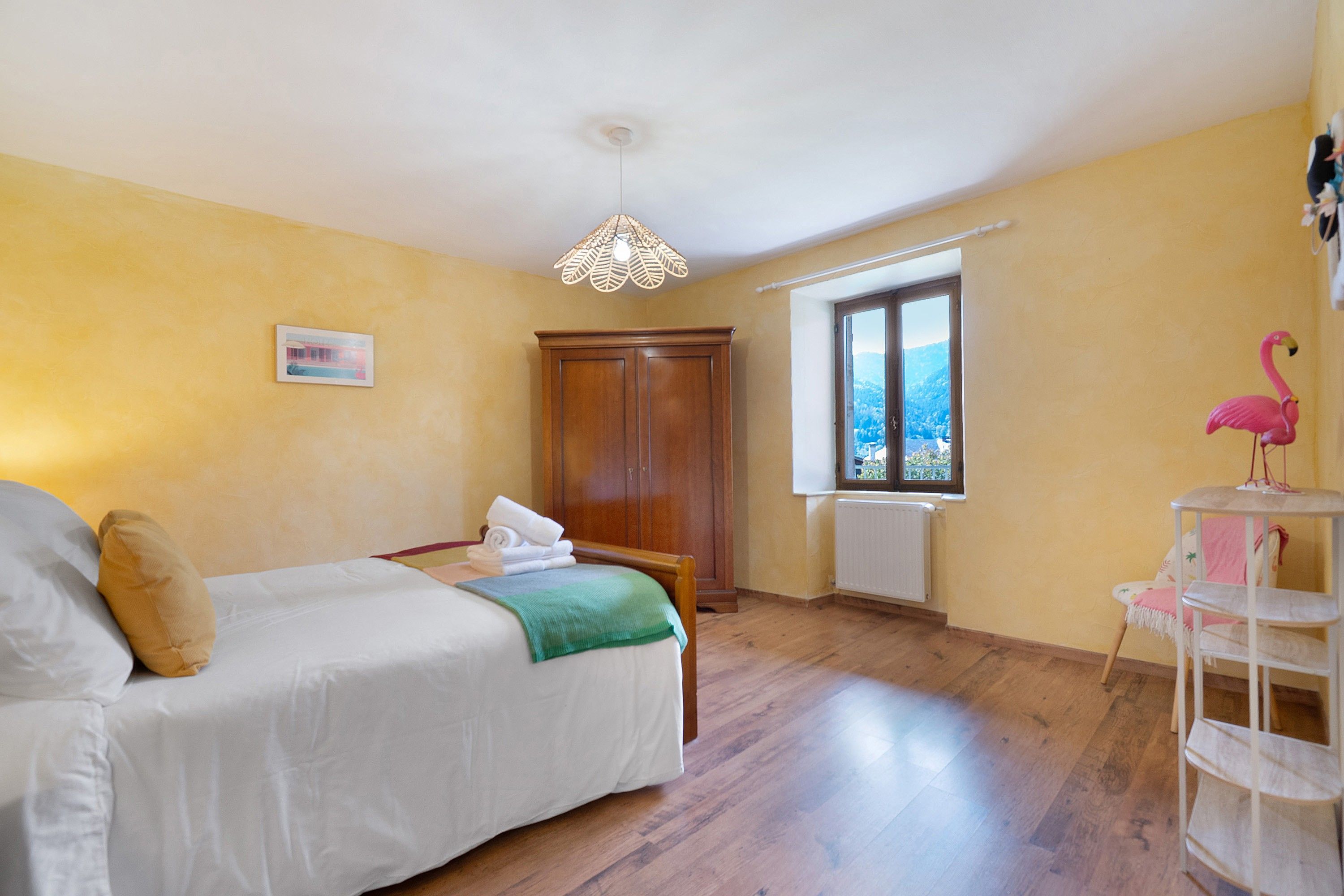 Une chambre à coucher avec un lit, une armoire en bois, une fenêtre donnant sur un beau paysage et un objet de décoration représentant un flamant rose.