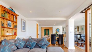 A modern living room with a grey sectional sofa, wooden shelves with various items, a dining area with a table and chairs, and a kitchen in the background. - 2 Bed in Redruth (oc-sanap) (Portreath)