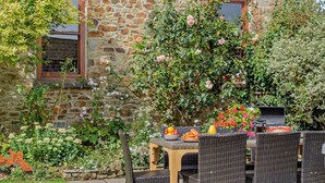 An outdoor patio with wicker furniture, a table with fruits and a beverage, and a stone building in the background. - 4 Bed in Bude (oc-ssham) (Stratton)
