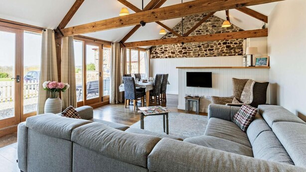 A spacious living area with a stone wall, wooden beams, a sofa set, a dining table, and a TV. - 3 Bed in Umberleigh (oc-09092) (Umberleigh)