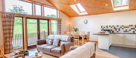 A cozy living room with a sofa, a coffee table, and a dining area with chairs and a table. There is a kitchen with a tiled backsplash and cabinets. Large windows provide a view of the outdoors.
