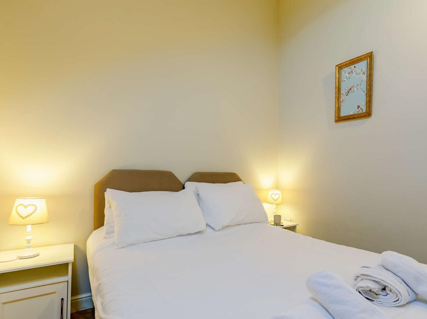 A hotel room with a bed, two bedside lamps, a heart-shaped lamp, and a framed picture on the wall.