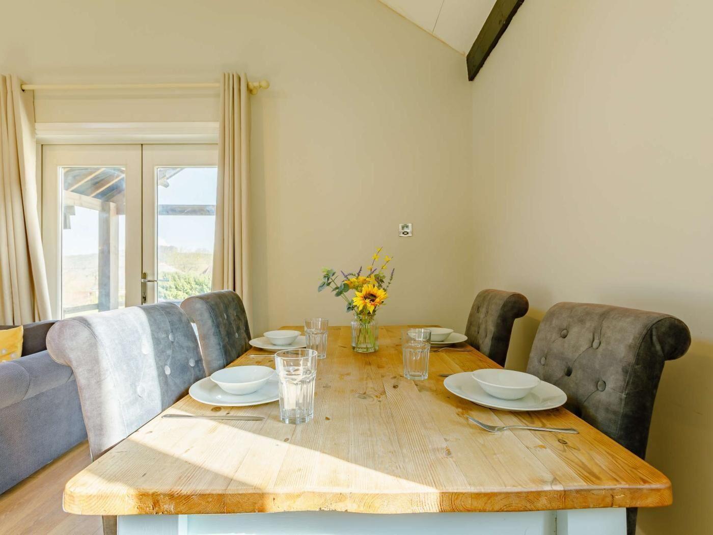 A dining area with a wooden table, four grey upholstered chairs, a vase with flowers, and a view through a glass door.
