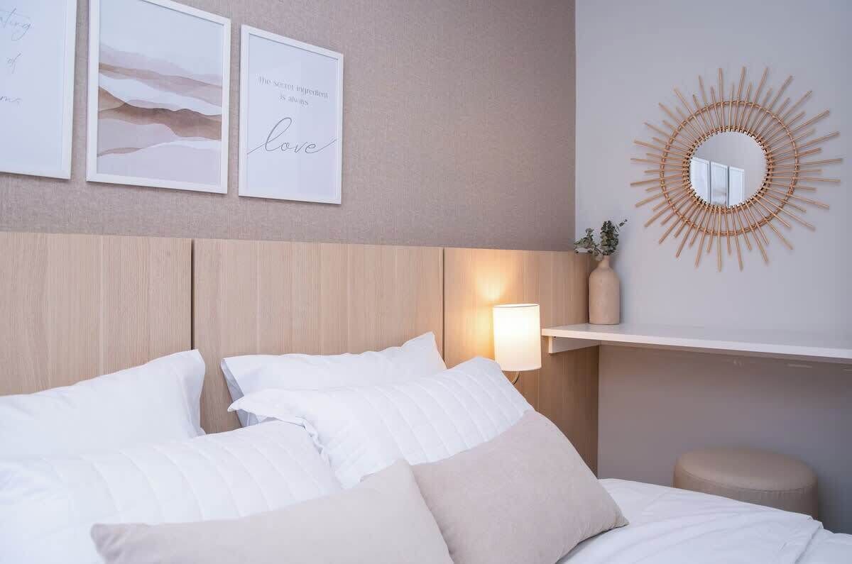 Un lit bien fait, avec une literie blanche et beige, un miroir fixé au mur et des œuvres d’art encadrées.
