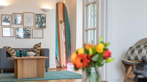 A living room with a sofa, coffee table, and a surfboard leaning against the wall. - 5 Bed in Bideford (oc-09156) (Northam)