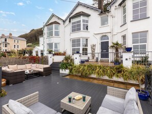Ein weißes Gebäude mit einer überdachten Terrasse, Sitzgelegenheiten im Freien und Blumentöpfen.