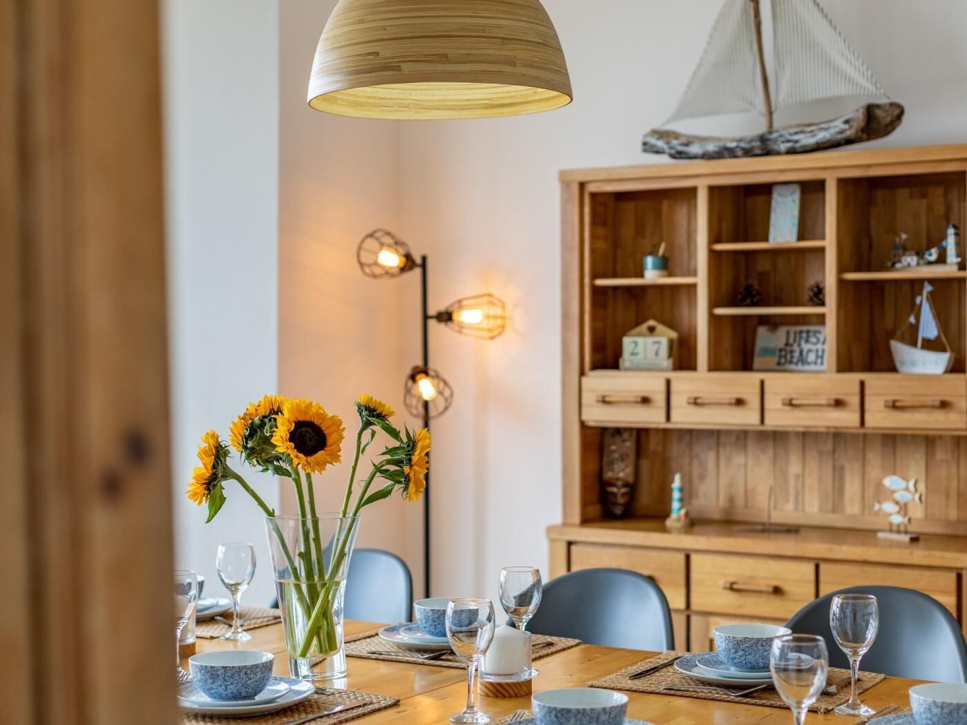A dining area with a wooden table set for four, surrounded by chairs. There is a vase with sunflowers on the table, and a wooden cabinet with shelves in the background.