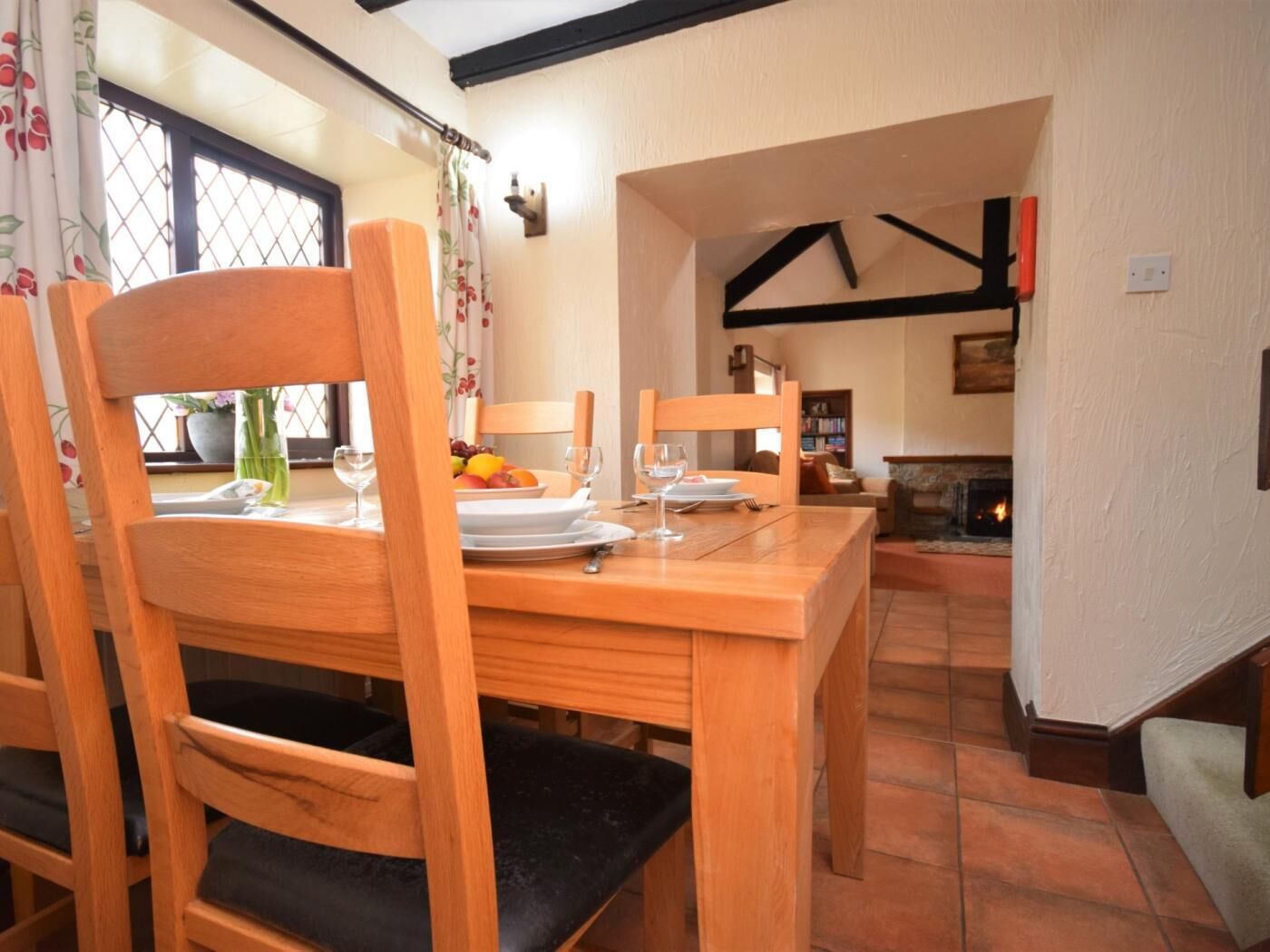 A dining area with wooden chairs and a table set for two, a fireplace in the background, and a window with curtains.