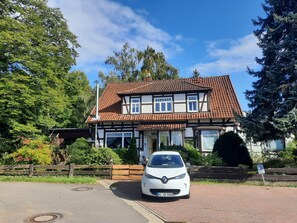 A two-story house with a tiled roof, a white car parked in front, and a wooden fence surrounding the property. - Apartment in a half-timbered house with a unique garden. Golf, Steinhuder Meer. (Rehburg-Loccum)