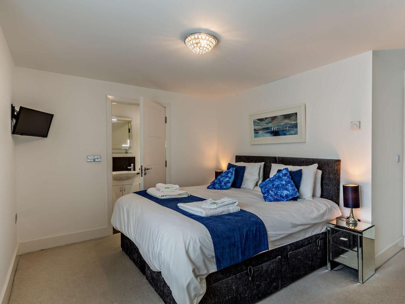 Een slaapkamer met een bed, nachtkastje, lamp en een aan de muur bevestigde televisie.