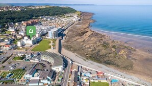 An aerial view of a coastal town with a beach, a large hotel with a curved roof, and a green speech bubble icon. - 2 Bed in Bideford (oc-sscpe) (Westward Ho!)