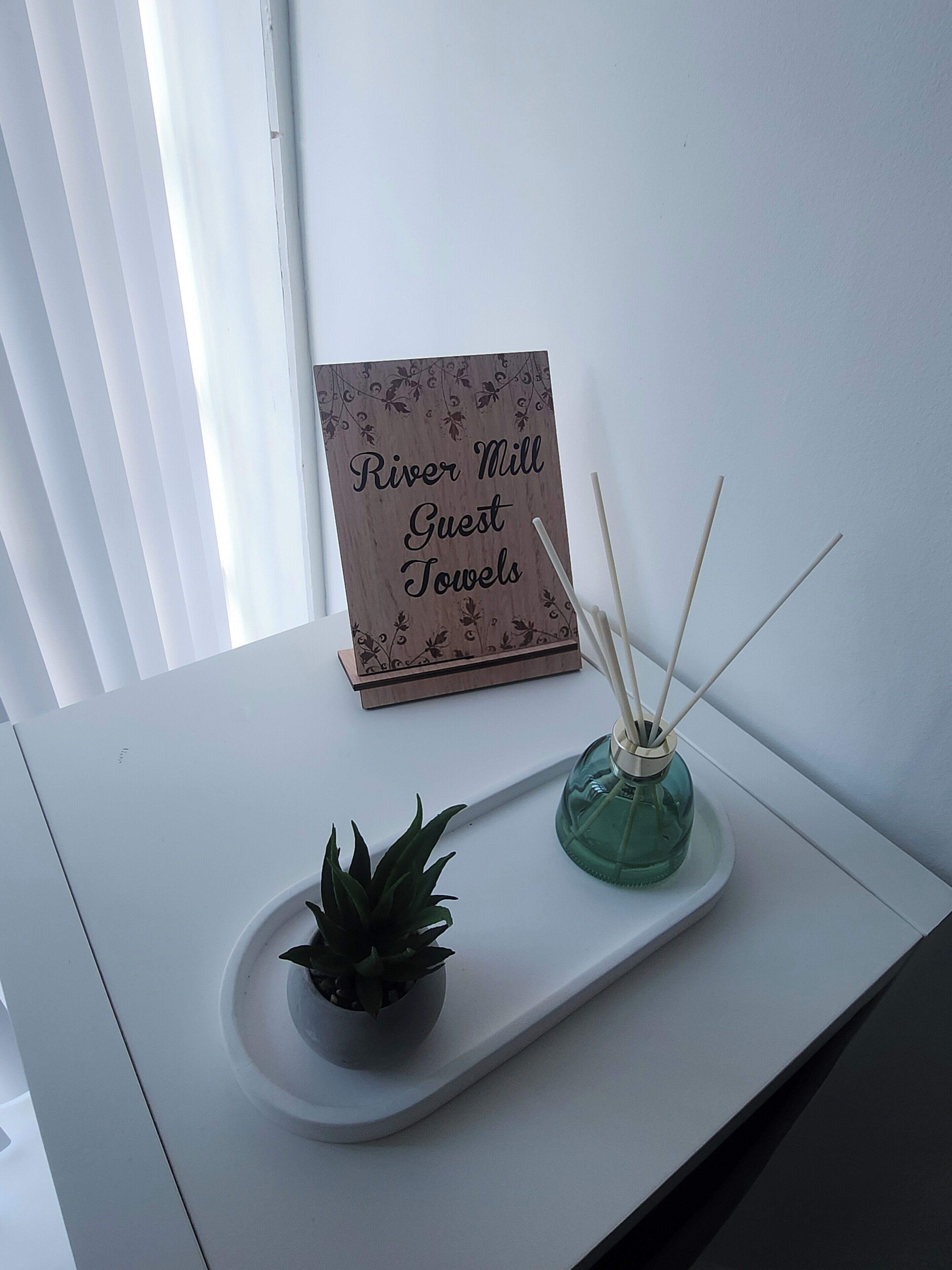 Ein weißer Tisch mit einem Schild, auf dem 'River Mill Guest Towels' steht, einer kleinen Topfpflanze und einem Schilferdiffusor.