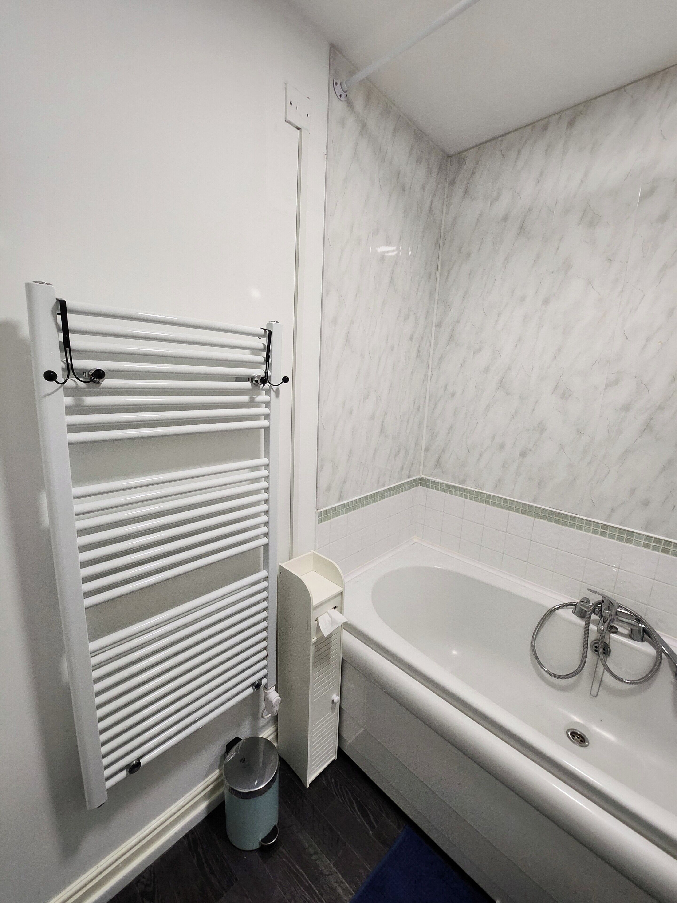A bathroom with a white bathtub, a towel rack, a waste bin, and a tiled wall.