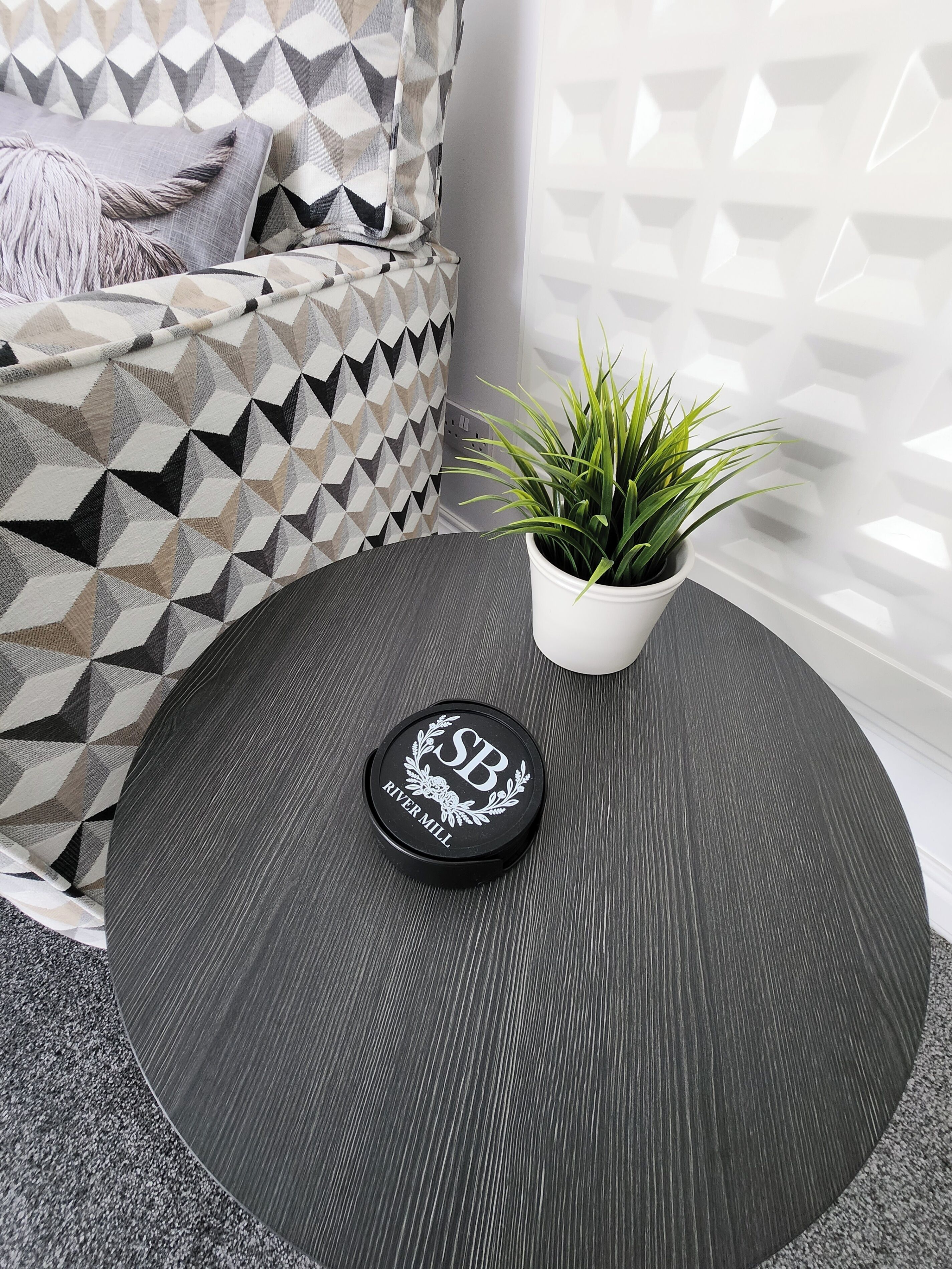 Een ronde tafel met een zwart oppervlak, een witte plant in pot en een zwart object met de tekst 'SB' en 'RIVER MILL'.