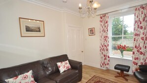 A living room with a brown leather sofa, a patterned rug, a small table, and a window with floral curtains. - 2 Bed in Huntly (oc-ab171) (Huntly)