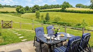 Un área de comedor al aire libre con una mesa lista para cuatro personas, rodeada de sillas y vista a un paisaje verde con árboles y una cerca.