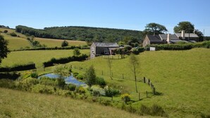 Paisaje rural con una casa pequeña, un estanque y colinas ondulantes al fondo.