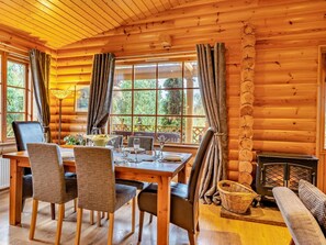 A wooden cabin interior with a dining table set for four, chairs, a sofa, a fireplace, and large windows with curtains.
