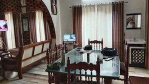 A dining area with a glass table, wooden chairs, a television, and a kitchen area with a sink and mirror. - Green Nestle Farmhouse  (Vaikom)