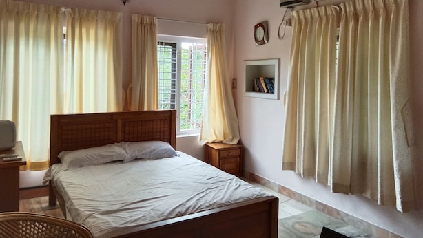 A bedroom with a bed, a bedside table, a nightstand, a wall-mounted air conditioner, a window with curtains, and a small shelf with books. - Green Nestle Farmhouse  (Vaikom)
