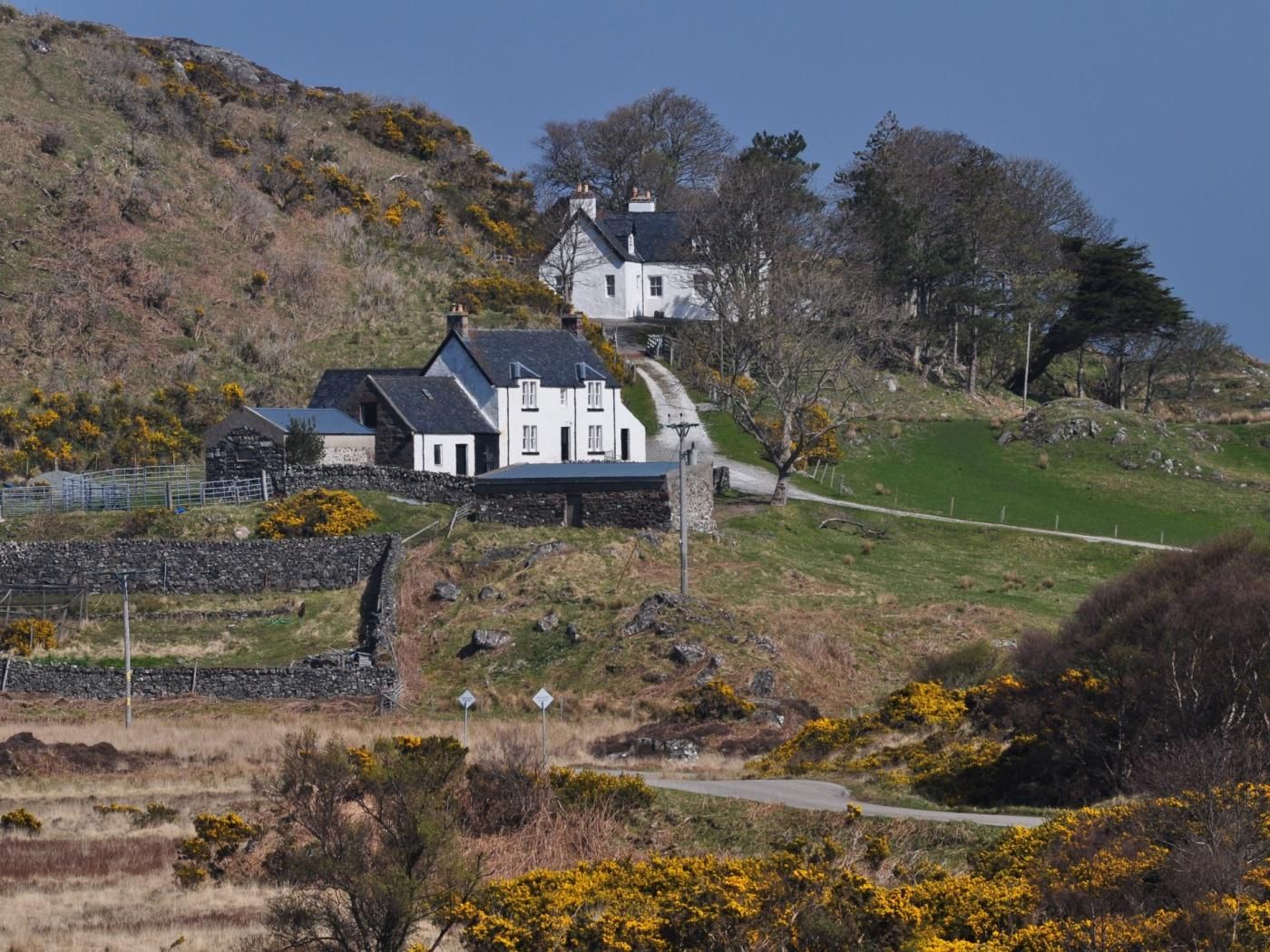 A white house with a black roof, surrounded by a stone wall and a grassy hill.
