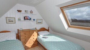 Ein Schlafzimmer mit zwei Einzelbetten, einer Holzkommode, einer kleinen Ablage mit Büchern und Dekoration sowie einem Fenster mit Blick ins Freie.