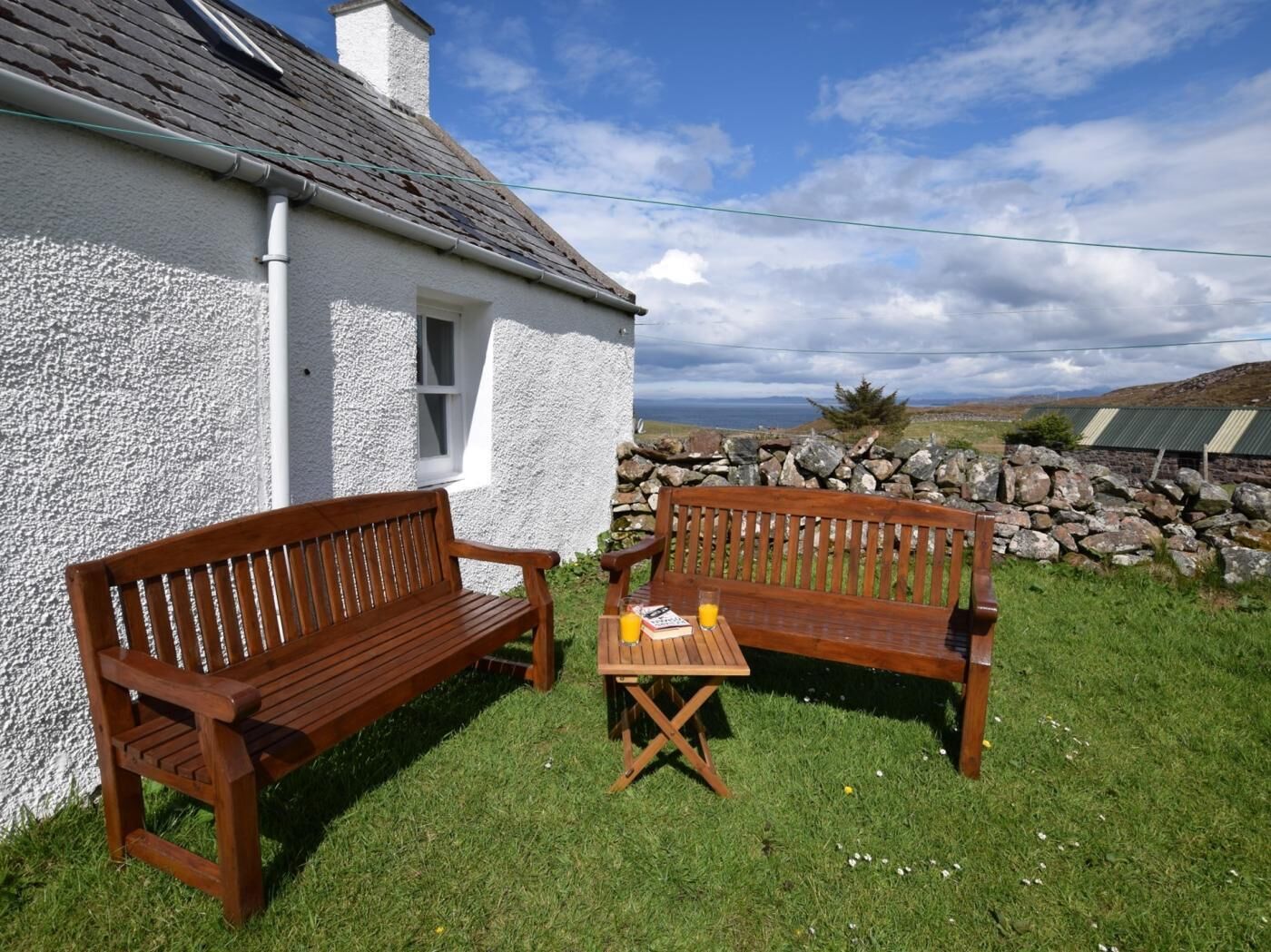 A white house with a stone wall, two wooden benches, and a table with a glass of orange juice.