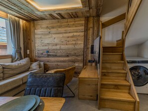 A cozy room with a wooden wall, a sofa, a chair, a small table, and a wooden staircase leading to a laundry area. - Modern apartment in Belle Plagne - 6 guests (Belle Plagne)