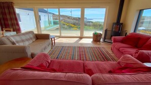 A living room with a sofa, a red couch, a fireplace, and a view of the sea. - 3 Bed in Lairg (oc-su164) (Scourie More)
