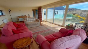 A spacious living area with a large window, a sofa, a chair, and a small table. - 3 Bed in Lairg (oc-su164) (Scourie More)