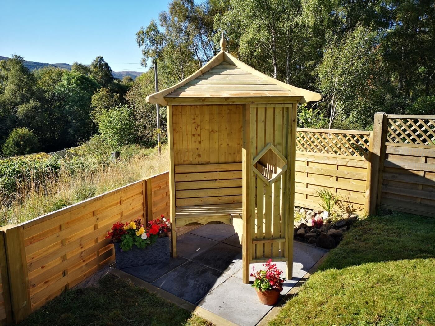 A wooden gazebo with a sloped roof, surrounded by a wooden fence, and featuring a small entrance with a decorative door.