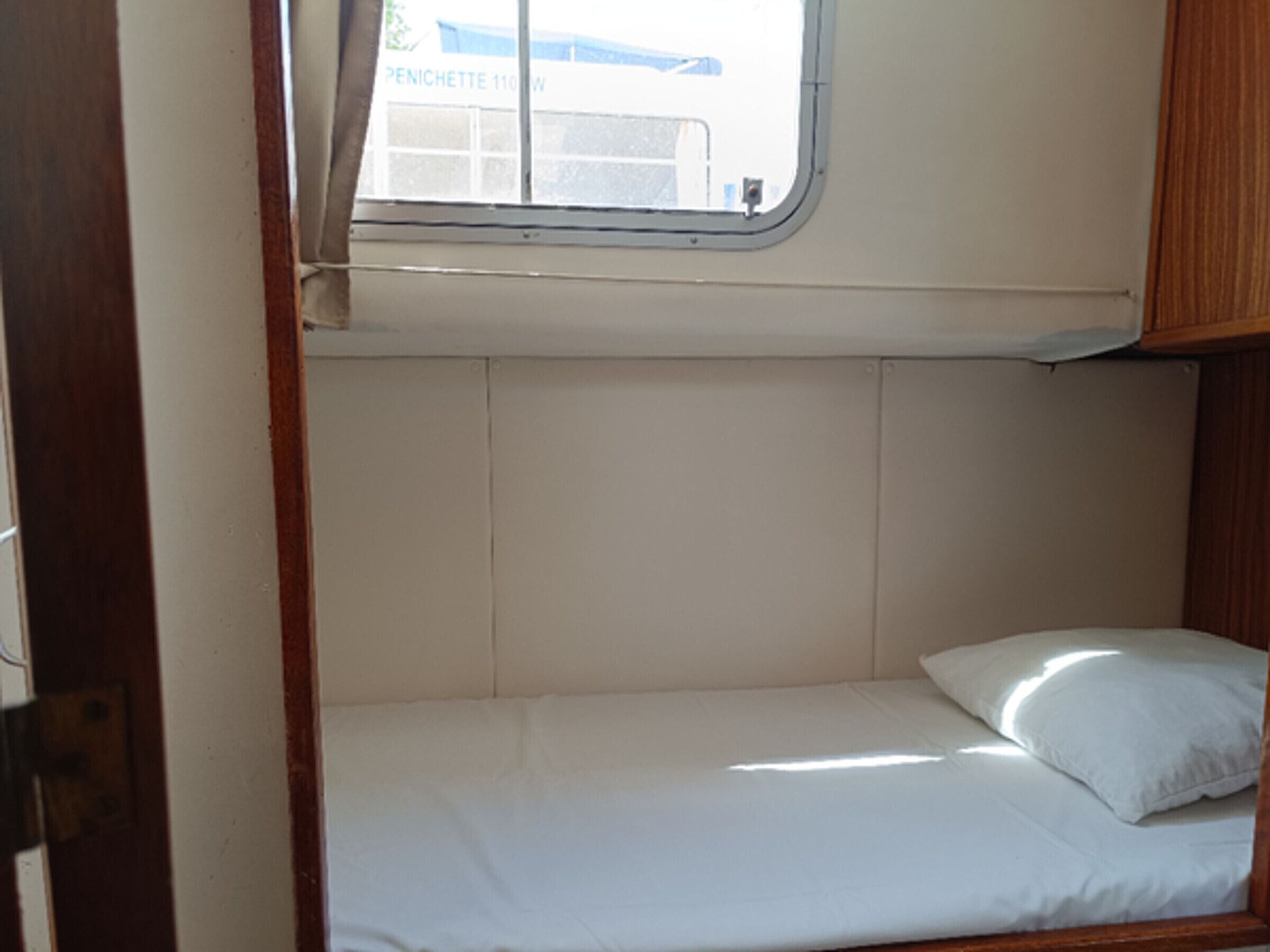 A small, single-bed cabin with a window, wooden headboard, and a white mattress.
