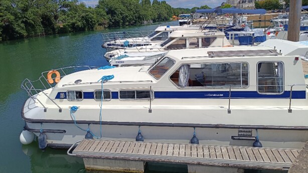 Ein Yachthafen mit mehreren angestaubten Booten, einer Holzsteganlage und einem Fluss mit Bäumen an den Ufern.