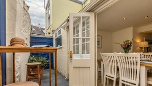 A cozy patio with a wooden table and chairs, a door leading to an interior dining area, and a blue gate in the background. - 3 Bed in Bideford (oc-insbr) (Instow)