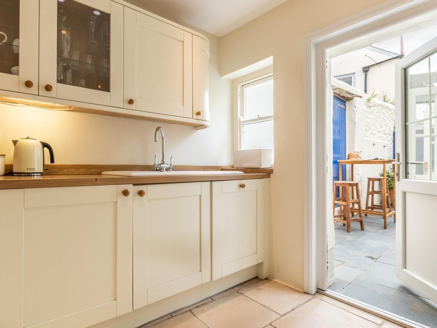 Een keuken met witte kastjes, een spoelbak en een waterkoker. Er is een raam en een deur naar een buitenruimte met een tafel en stoelen.