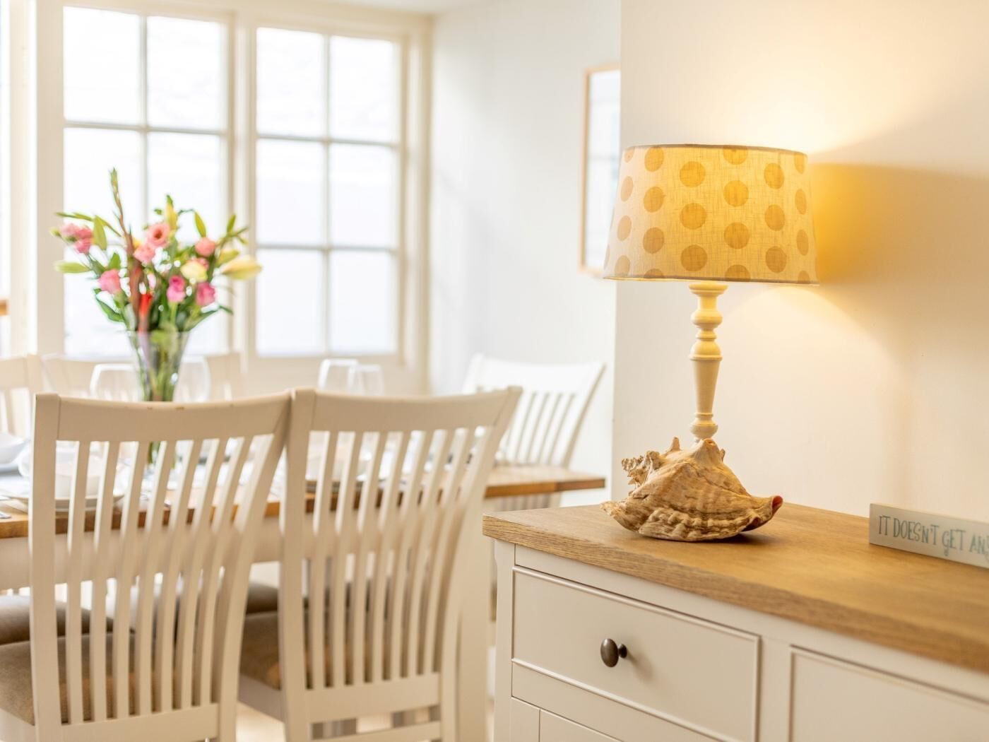 Un coin repas avec des chaises blanches, une table, une lampe avec un abat-jour à pois et un vase rempli de fleurs.